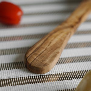 Puede incluir: Primer plano de un cuchillo de mantequilla de madera con un patr&oacute;n de veta de madera natural. El cuchillo est&aacute; sobre una superficie rayada blanca y marr&oacute;n.