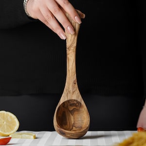 Puede incluir: Un cuchar&oacute;n de madera de olivo con un patr&oacute;n de veta de madera natural. El cuchar&oacute;n es grande y tiene un cuenco redondeado.