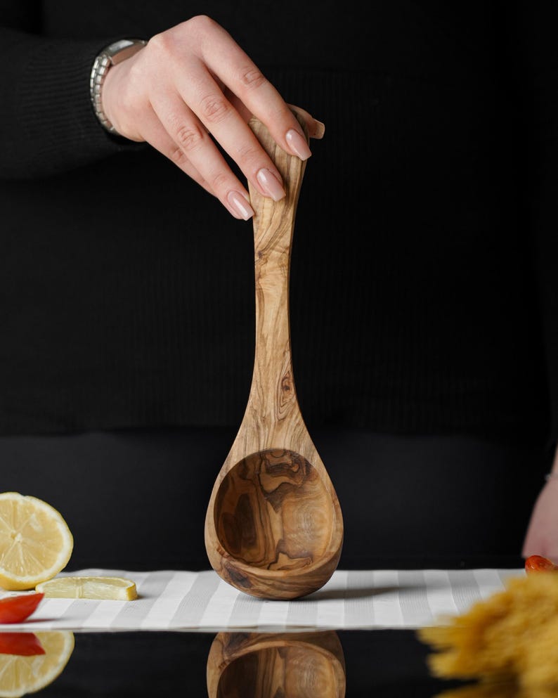 Puede incluir: Un cuchar&oacute;n de madera de olivo con un patr&oacute;n de veta de madera natural. El cuchar&oacute;n es grande y tiene un cuenco redondeado.