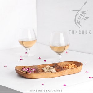 Könnte beinhalten: Ein Serviertablett aus Olivenholz mit drei Fächern. Das Tablett ist gefüllt mit Pistazien, getrockneten Rosenblüten und gebratenen Erdnüssen. Im Hintergrund stehen zwei Gläser Weißwein. Das Tablett ist handgefertigt und hat eine natürliche Holzoptik. Der Text "Tunsouk" und "Handcrafted Olivewood" ist auf dem Bild zu sehen.
