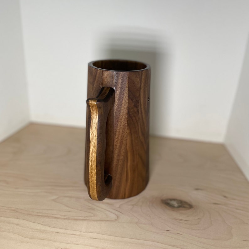 May include: A handcrafted wooden beer stein with a dark brown finish. The stein features a large, sturdy handle with a lighter wood accent. The body of the stein has a faceted design. The mug is empty and sits on a light-colored wooden surface.