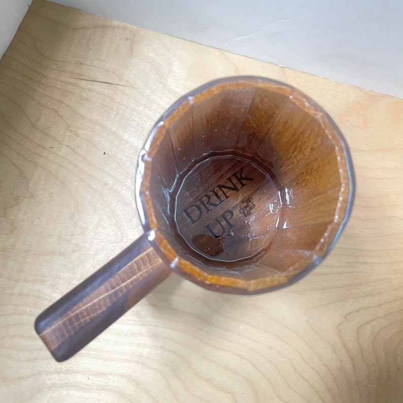 May include: A wooden mug with a handle, featuring a light brown exterior and a darker brown handle. The words "DRINK UP" are printed on the bottom of the mug. The mug is empty.