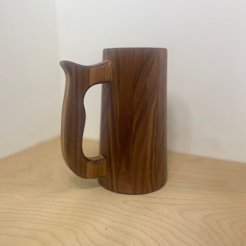 May include: A handcrafted wooden beer mug with a curved handle. The mug is made of dark brown wood and has a cylindrical shape. The handle is also made of wood and is attached to the side of the mug.