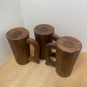 May include: Four handcrafted wooden mugs with a dark brown finish. Each mug features a unique handle design and a circular top with engraved text and a mountain and tree logo. The mugs are displayed on a light wood surface.