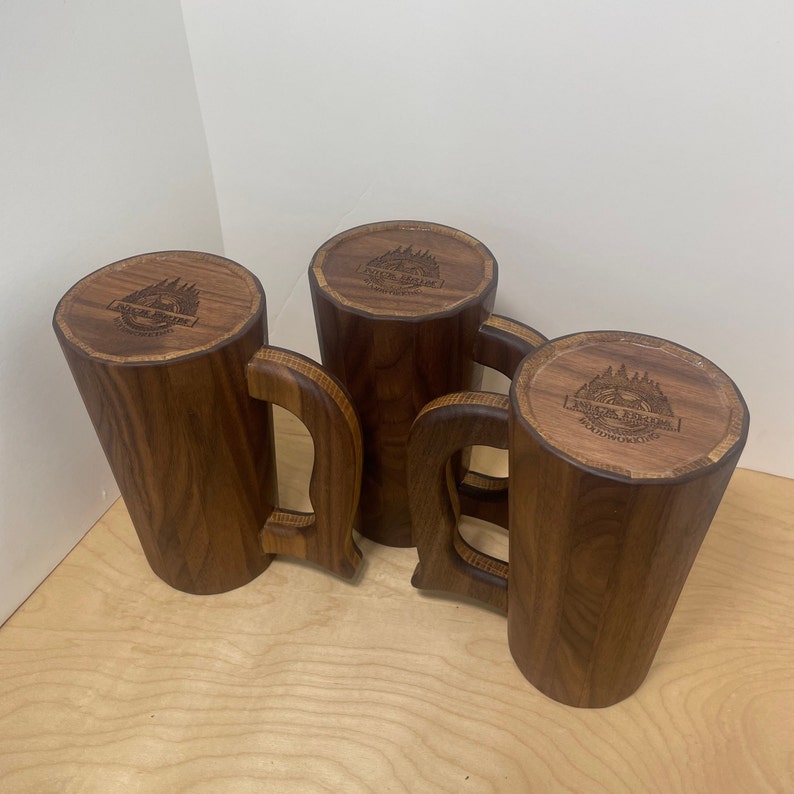 May include: Four handcrafted wooden mugs with a dark brown finish. Each mug features a unique handle design and a circular top with engraved text and a mountain and tree logo. The mugs are displayed on a light wood surface.