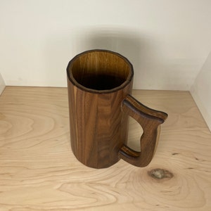 May include: A handcrafted wooden mug with a large handle. The mug is made of dark brown wood and has a cylindrical shape. The handle is curved and attached to the side of the mug. The mug is designed for beverages.