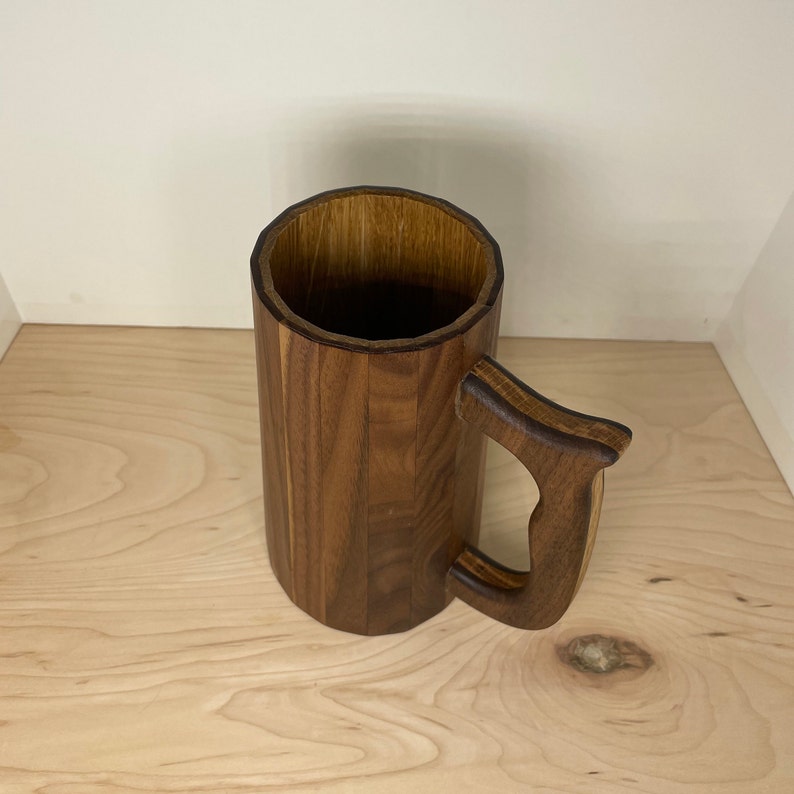 May include: A handcrafted wooden mug with a large handle. The mug is made of dark brown wood and has a cylindrical shape. The handle is curved and attached to the side of the mug. The mug is designed for beverages.