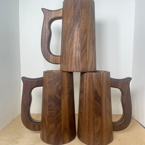 May include: Three wooden beer mugs stacked. Each mug is a dark brown color with a large handle. The mugs are made of vertical wooden planks, giving them a rustic appearance. The mugs are empty and ready to be filled.