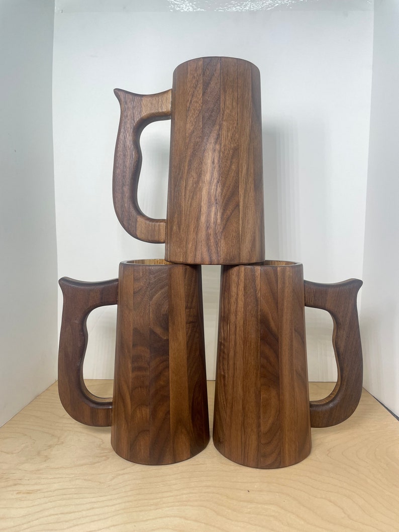 May include: Three wooden beer mugs stacked. Each mug is a dark brown color with a large handle. The mugs are made of vertical wooden planks, giving them a rustic appearance. The mugs are empty and ready to be filled.