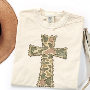May include: Cream-colored t-shirt featuring a large, embroidered cross. The cross design incorporates a camouflage pattern in shades of green, brown, and tan. The shirt has a crew neck and a tag that reads "Comfort Colors".