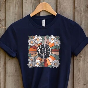 May include: Navy blue t-shirt with a retro design. The graphic features the text "HERE COMES THE SUN" in a circular design with orange, brown, and tan rays, surrounded by white daisies. The shirt is hanging on a wooden hanger.
