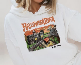 May include: White hoodie with a colorful graphic featuring the text "Halloweentown" above an illustration of a town scene with a pumpkin and buildings. The text "Est. 1998" is also included in the design.