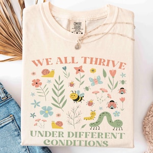 May include: Cream-colored t-shirt with the text "WE ALL THRIVE UNDER DIFFERENT CONDITIONS" and a colorful graphic of flowers, insects, and a caterpillar. The shirt is folded next to blue jeans, white sneakers, and a beige blanket.