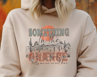 May include: A beige hoodie with a graphic print of a desert scene with cacti and the text "Something in the Orange tells me we're not done".