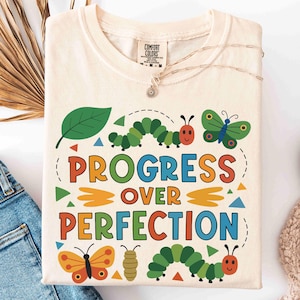 May include: Cream-colored t-shirt with the words "PROGRESS OVER PERFECTION" in colorful letters. The shirt features cartoon caterpillars, butterflies, and a leaf design. The shirt is folded and placed on a white surface with blue jeans and white shoes.