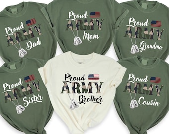 Op de afbeelding: Zes groene en één crèmekleurige T-shirts met een camouflagepatroon en de woorden "Proud Army" met een kleine Amerikaanse vlag en een hondenpenning. De shirts hebben verschillende woorden erop, waaronder "Papa", "Mama", "Oma", "Zus", "Broer" en "Neef".