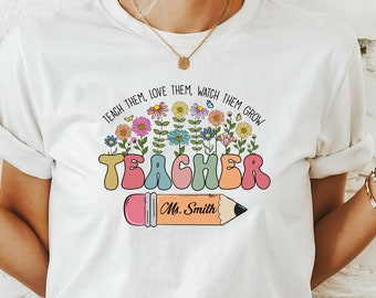 May include: White t-shirt with a colorful graphic design for teachers. The design includes the word "TEACHER" in bubble letters, a pencil, and the phrase "TEACH THEM, LOVE THEM, WATCH THEM GROW".