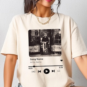 May include: A white t-shirt with a black and white image of a person playing a guitar. The image is framed with a black border and has a play button, rewind button, and fast forward button below it. The text "Song Name" and "Artist Name" are below the image. The text "0:20" and "3:35" are at the bottom left and right corners of the image.
