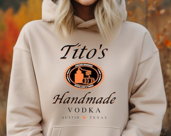 May include: A beige hoodie with a black and orange logo that says "Tito's Handmade Vodka Austin * Texas". The logo features a stylized image of a bottle and barrels.
