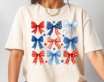 May include: A cream-colored t-shirt featuring a pattern of nine bows in red, white, and blue. The bows are arranged in three rows of three, with stars interspersed between them. The shirt has short sleeves and a relaxed fit.