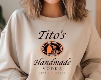 May include: A beige sweatshirt with a graphic design featuring the text "Tito's Handmade Vodka" and a logo with the text "Award Winning Distilled 5 Times".