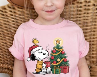 May include: A pink t-shirt featuring a cartoon Snoopy character wearing a Santa hat and holding a mug, with a Christmas tree and presents. The shirt is worn by a young child wearing a brown hat.