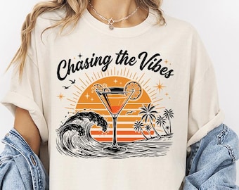 Può includere: Una t-shirt color crema con una stampa grafica raffigurante un bicchiere da cocktail, un grande sole arancione, palme e onde del mare. Il testo 'Chasing the Vibes' è stampato in nero sopra la grafica, che include anche piccole stelle e uccelli.