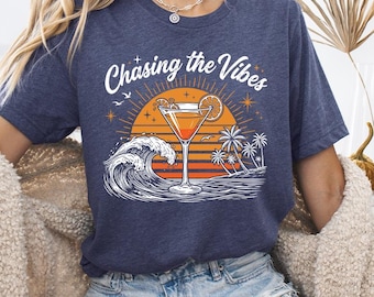 Può includere: Una t-shirt a maniche corte blu navy mélange con una stampa grafica raffigurante un bicchiere da cocktail, una grande onda oceanica, palme e un tramonto. Il testo "Chasing the Vibes" è stampato in bianco sopra la grafica.