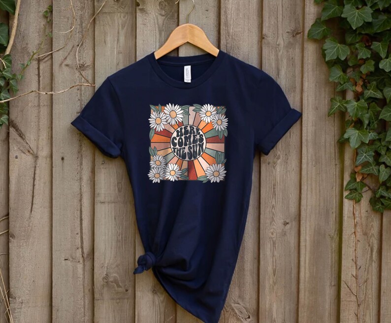 May include: Navy blue t-shirt with a retro design. The graphic features the text "HERE COMES THE SUN" in a circular design with orange, brown, and tan rays, surrounded by white daisies. The shirt is hanging on a wooden hanger.