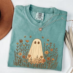 May include: A teal Comfort Colors t-shirt featuring a ghost design surrounded by flowers and bees. The ghost is beige, and the flowers are orange and yellow. A silver necklace with a small pendant is draped over the shirt.