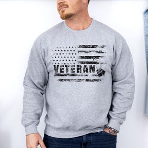 Peut inclure: Un sweat-shirt gris avec un motif de drapeau am&eacute;ricain vieilli et le mot "Veteran" imprim&eacute; en noir. Le drapeau a une rang&eacute;e d'&eacute;toiles en haut et une plaque d'identit&eacute; pendante en bas.
