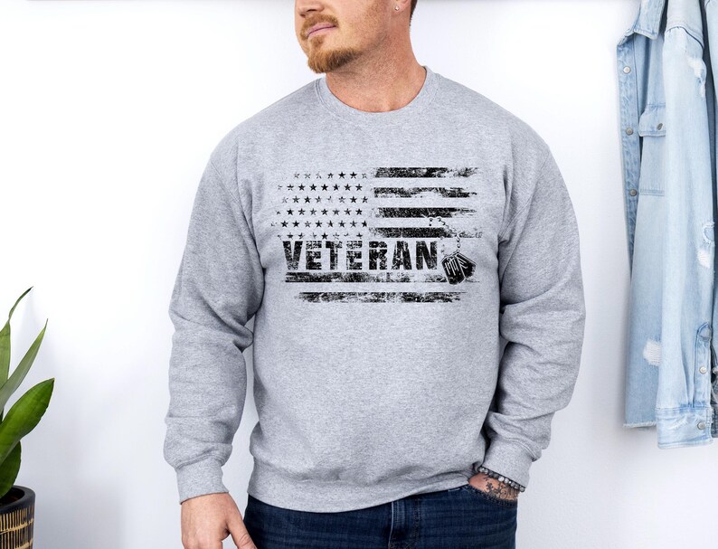 Peut inclure: Un sweat-shirt gris avec un motif de drapeau am&eacute;ricain vieilli et le mot "Veteran" imprim&eacute; en noir. Le drapeau a une rang&eacute;e d'&eacute;toiles en haut et une plaque d'identit&eacute; pendante en bas.