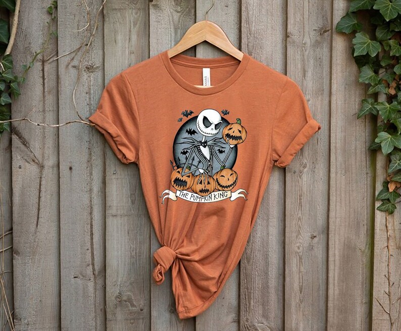 May include: Orange t-shirt featuring a graphic of Jack Skellington from The Nightmare Before Christmas, holding a pumpkin. The design includes pumpkins, bats, and the text "THE PUMPKIN KING" on a banner. The shirt is hanging on a wooden hanger.