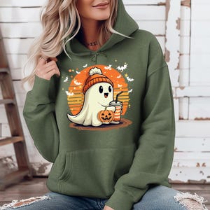 May include: A green hooded sweatshirt featuring a cute cartoon ghost wearing a hat and holding a pumpkin and a drink. The ghost is in front of a sunset with bats flying around it.
