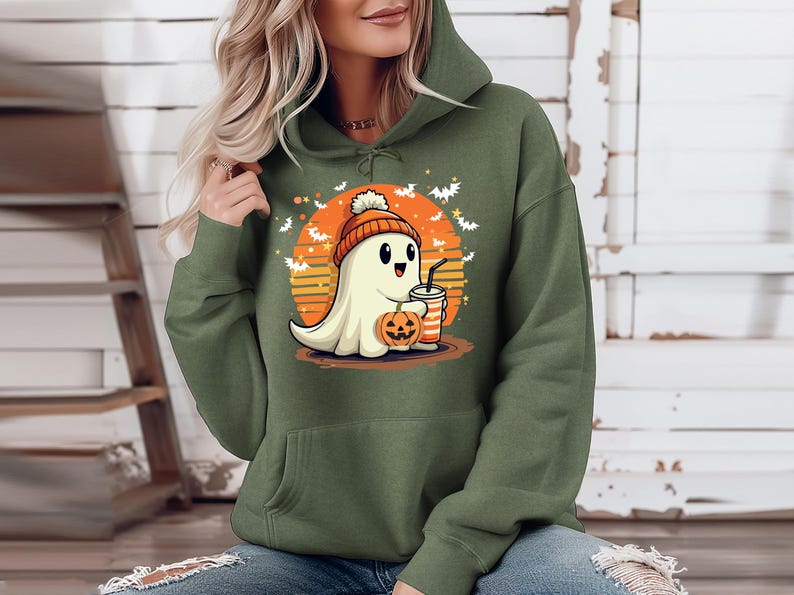 May include: A green hooded sweatshirt featuring a cute cartoon ghost wearing a hat and holding a pumpkin and a drink. The ghost is in front of a sunset with bats flying around it.