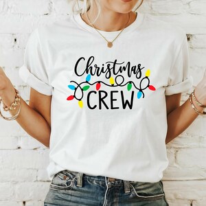 Puede incluir: Camiseta blanca con la frase "Christmas CREW" en letras negras, adornada con una cadena de luces navide&ntilde;as de colores. La camiseta es de estilo casual, con cuello redondo.