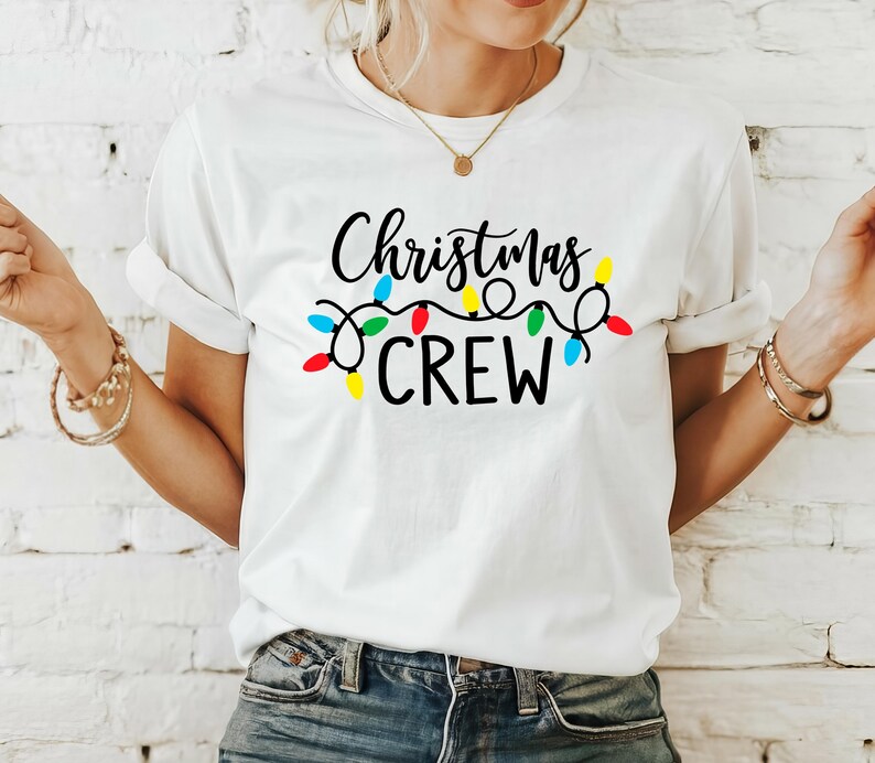 Puede incluir: Camiseta blanca con la frase "Christmas CREW" en letras negras, adornada con una cadena de luces navide&ntilde;as de colores. La camiseta es de estilo casual, con cuello redondo.