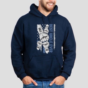 May include: Navy blue hooded sweatshirt with a graphic of five fists with the names "Lily", "Uncle", "Saben", "Jack", and "Best Uncle Ever" printed on the fists. The graphic is on a blue and white American flag background.