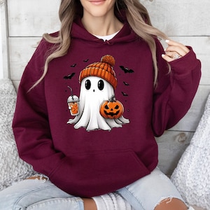 Pu&ograve; includere: Felpa con cappuccio bordeaux con un fantasma dei cartoni animati con un berretto arancione, una zucca e una bevanda. Pipistrelli neri volano intorno al fantasma. Il fantasma ha grandi occhi neri e un lenzuolo bianco.
