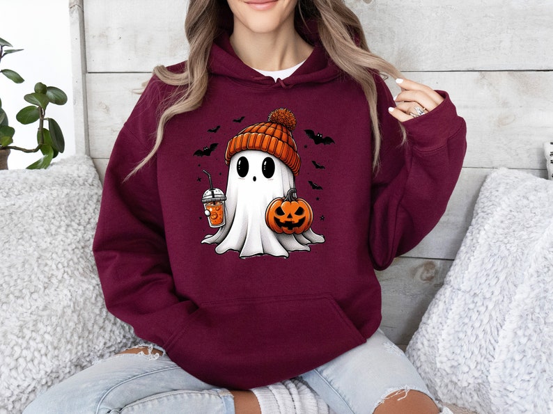Pu&ograve; includere: Felpa con cappuccio bordeaux con un fantasma dei cartoni animati con un berretto arancione, una zucca e una bevanda. Pipistrelli neri volano intorno al fantasma. Il fantasma ha grandi occhi neri e un lenzuolo bianco.