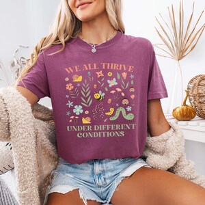 May include: A maroon t-shirt with the text "WE ALL THRIVE UNDER DIFFERENT CONDITIONS" in a square design with colorful illustrations of flowers, bees, and a caterpillar. The shirt is worn with denim shorts.