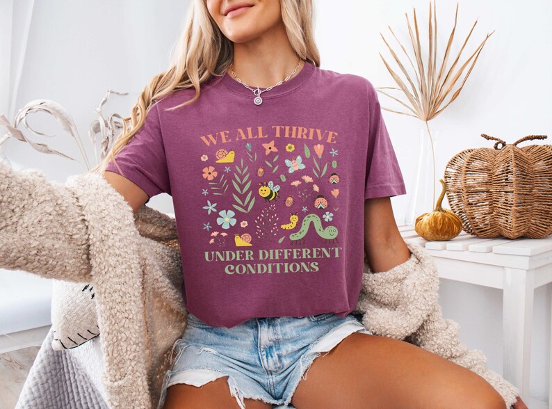 May include: A maroon t-shirt with the text "WE ALL THRIVE UNDER DIFFERENT CONDITIONS" in a square design with colorful illustrations of flowers, bees, and a caterpillar. The shirt is worn with denim shorts.