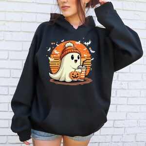 May include: Black hooded sweatshirt featuring a graphic of a ghost wearing a beanie and holding a pumpkin and a cup. The ghost is in front of a sunset with bats flying around it.