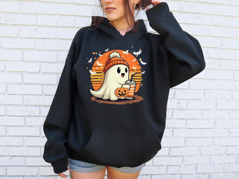 May include: Black hooded sweatshirt featuring a graphic of a ghost wearing a beanie and holding a pumpkin and a cup. The ghost is in front of a sunset with bats flying around it.