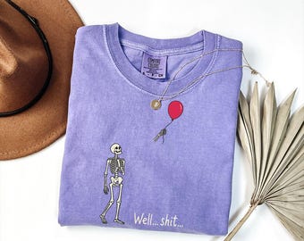 May include: A lavender short-sleeve t-shirt featuring a graphic of a skeleton holding a red balloon with the text "Well... shit..." printed below. The shirt has a Comfort Colors brand label at the inner neckline and a crew neck collar design.