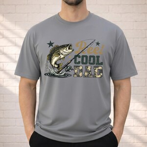 May include: Gray t-shirt featuring a graphic of a leaping fish, fishing rod, and the text "Reel Cool Dad" in a mix of fonts and colors. The word "DAD" is in a camouflage pattern. The shirt is short-sleeved.
