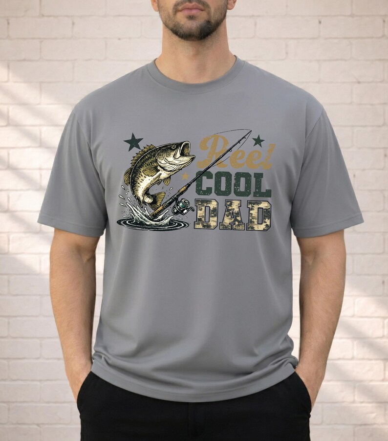 May include: Gray t-shirt featuring a graphic of a leaping fish, fishing rod, and the text "Reel Cool Dad" in a mix of fonts and colors. The word "DAD" is in a camouflage pattern. The shirt is short-sleeved.