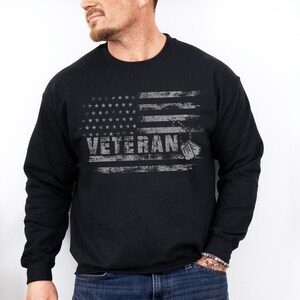 Peut inclure: Sweat-shirt noir avec un motif de drapeau am&eacute;ricain vieilli et le mot "Veteran" en lettres blanches. Deux m&eacute;dailles militaires pendent en bas du drapeau.