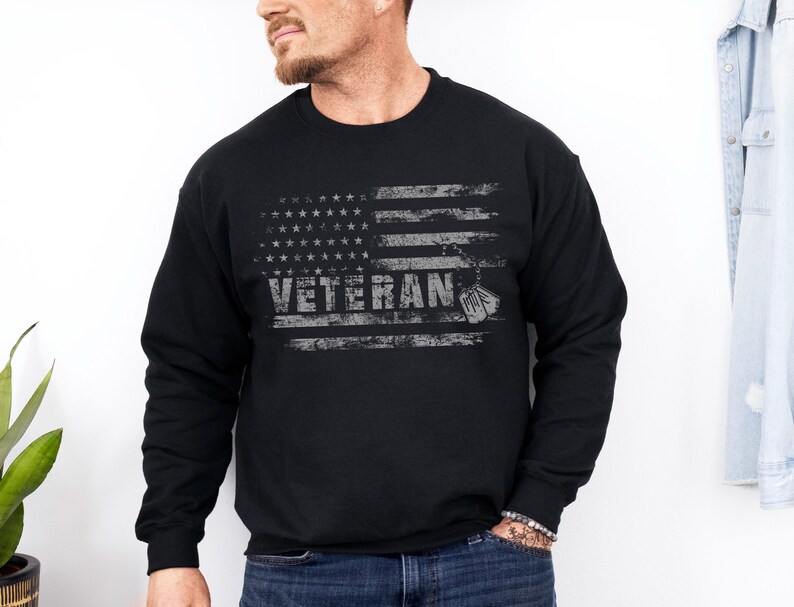 Peut inclure: Sweat-shirt noir avec un motif de drapeau am&eacute;ricain vieilli et le mot "Veteran" en lettres blanches. Deux m&eacute;dailles militaires pendent en bas du drapeau.