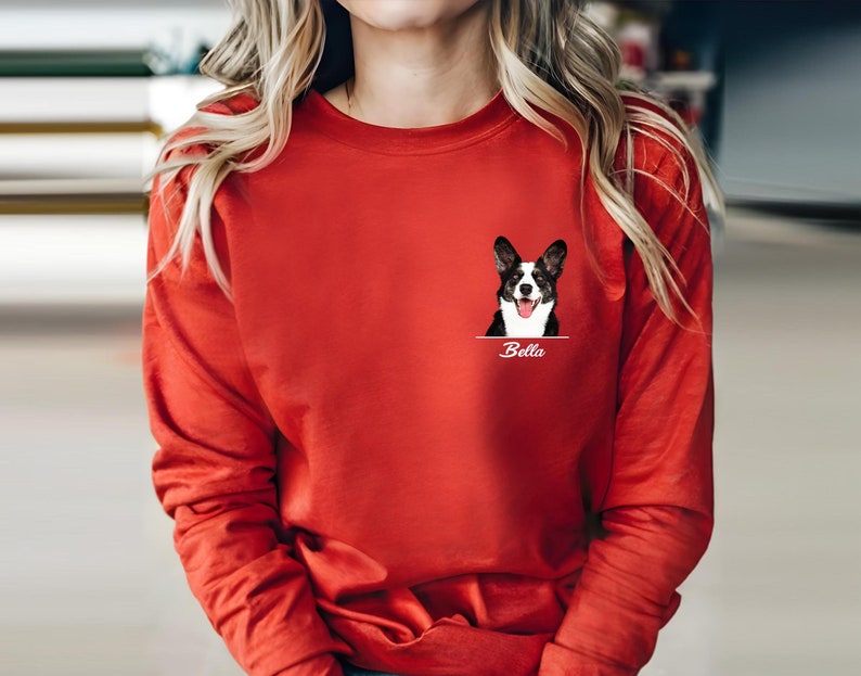 May include: A long-sleeved red shirt featuring a graphic of a dog with black and white markings and the name "Bella" underneath. The dog graphic is on the left chest area of the shirt.